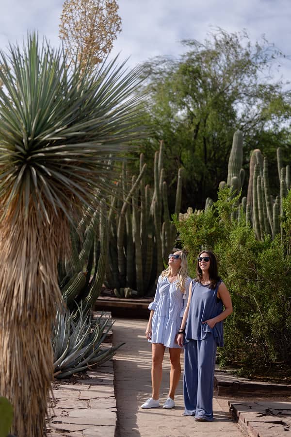 Two women pose on a stone path in a desert botanical garden, surrounded by tall cacti, yucca, and greenery under a bright sky.